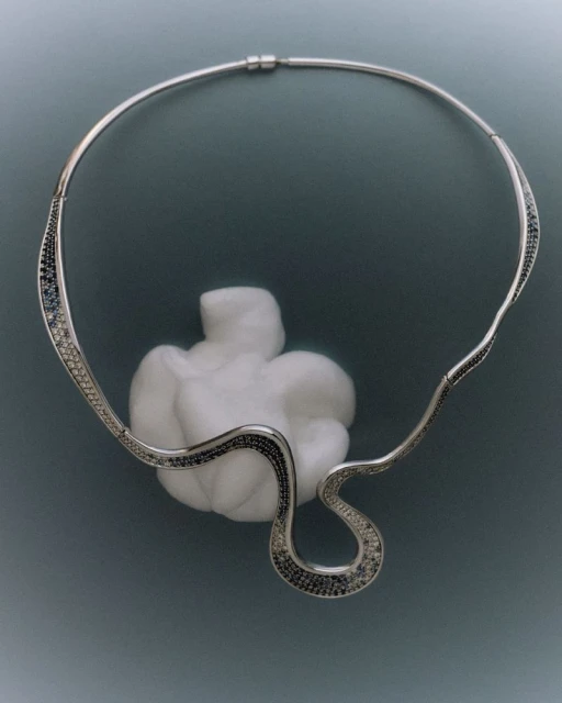 Silver metal necklace with embedded black stones and an open, curving design, displayed with a white textured form on a gray background.