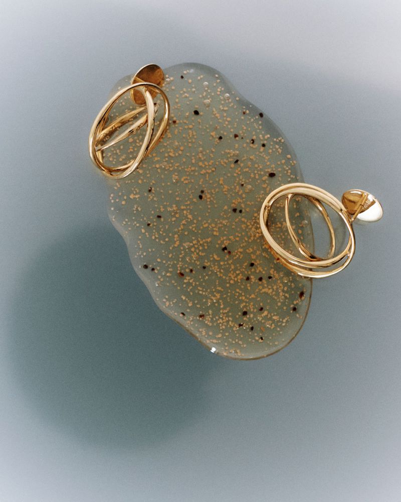 High-angle product shot of two gold earrings placed on a shimmering green gel-like surface against a muted light blue background.