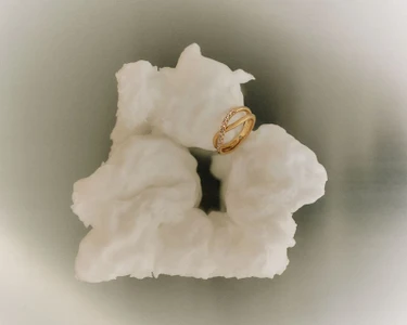 Gold double band ring with embedded crystals resting on a white sculptural rock formation against a soft grey gradient background.
