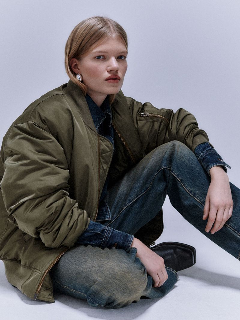 Model seated on the floor wearing an olive green bomber jacket over a denim shirt and blue jeans against a light background.