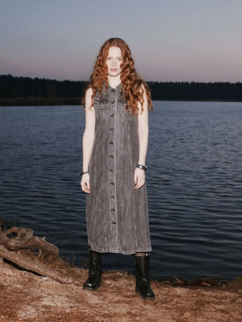 A woman with long red hair stands near a lake wearing a grey denim dress and black boots, creating a fashion-forward editorial style.