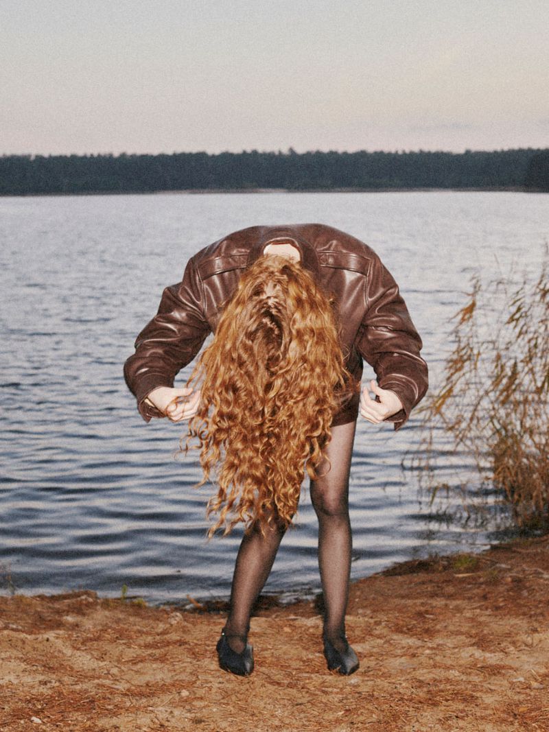 A woman in a leather jacket bends over on a lakeside shore with her wavy red hair cascading down, in a natural outdoor environment.