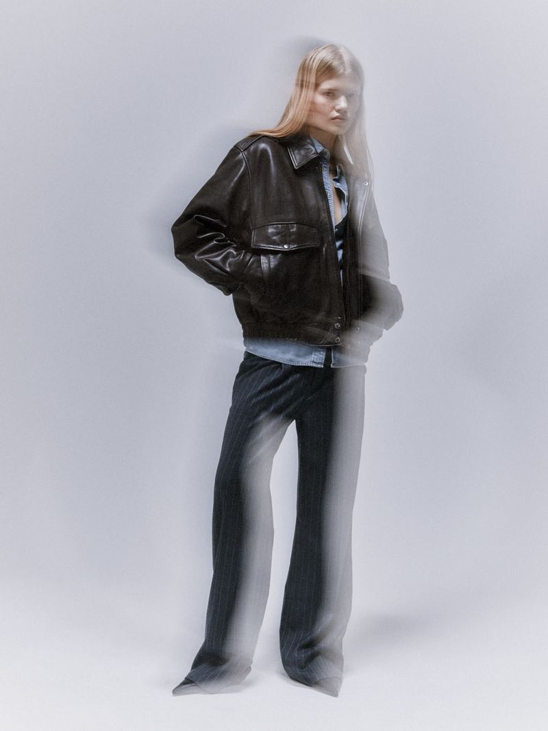 Model wearing a dark leather bomber jacket and denim shirt against a grey studio backdrop with motion blur effects.