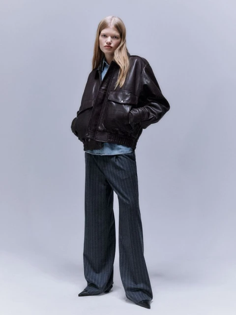 A fair-skinned woman poses in a dark leather bomber jacket and pinstripe pants against a solid grey studio backdrop.