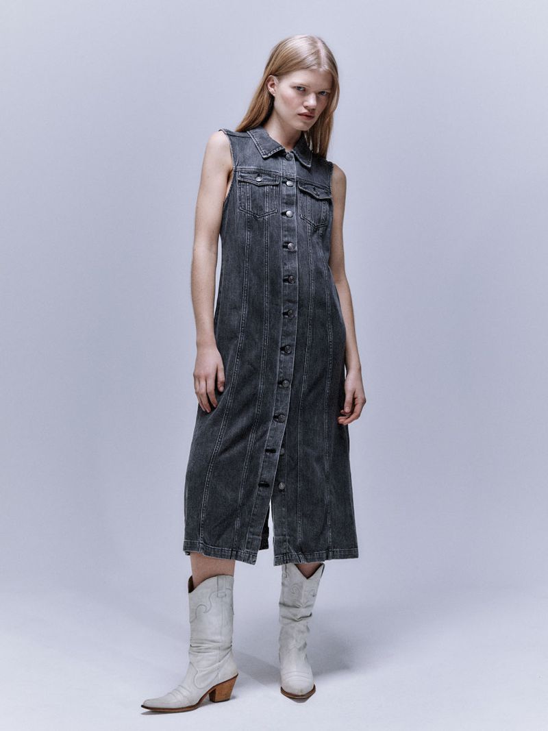 A fair-skinned woman poses in a sleeveless, ankle-length, grey denim dress and white Western boots in a bright, neutral studio setting.