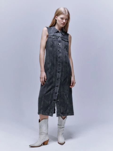 A fair-skinned woman poses in a sleeveless, ankle-length, grey denim dress and white Western boots in a bright, neutral studio setting.