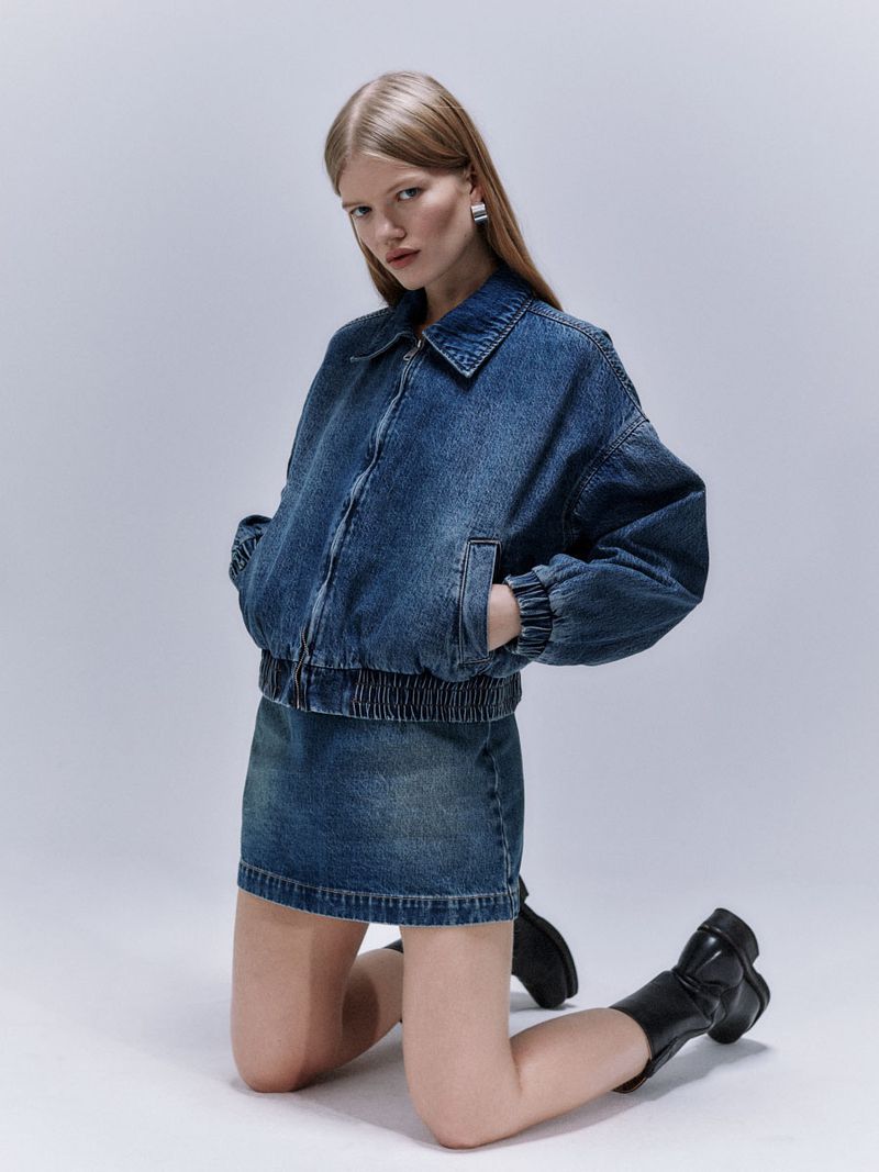 A woman with long blonde hair poses on her knees while wearing a denim jacket, a denim skirt, and black boots in a studio setting.