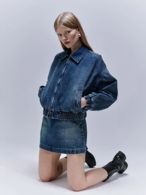 A woman with long blonde hair poses on her knees while wearing a denim jacket, a denim skirt, and black boots in a studio setting.