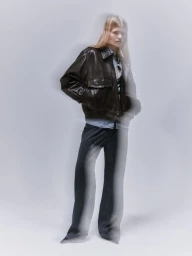 Model wearing a dark leather bomber jacket and denim shirt against a grey studio backdrop with motion blur effects.