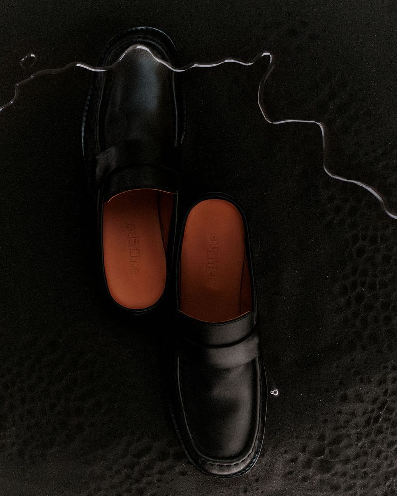 Studio product shot of a pair of black leather slip-on shoes on a black leather background, accented with a thin stream of water.
