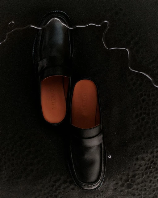 Studio product shot of a pair of black leather slip-on shoes on a black leather background, accented with a thin stream of water.