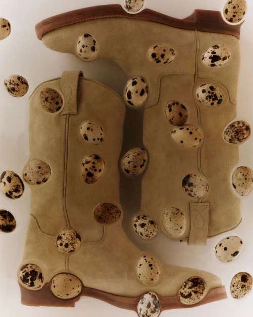 Pair of tan cowboy boots surrounded by speckled quail eggs in a studio shot on a white backdrop with diffused lighting.
