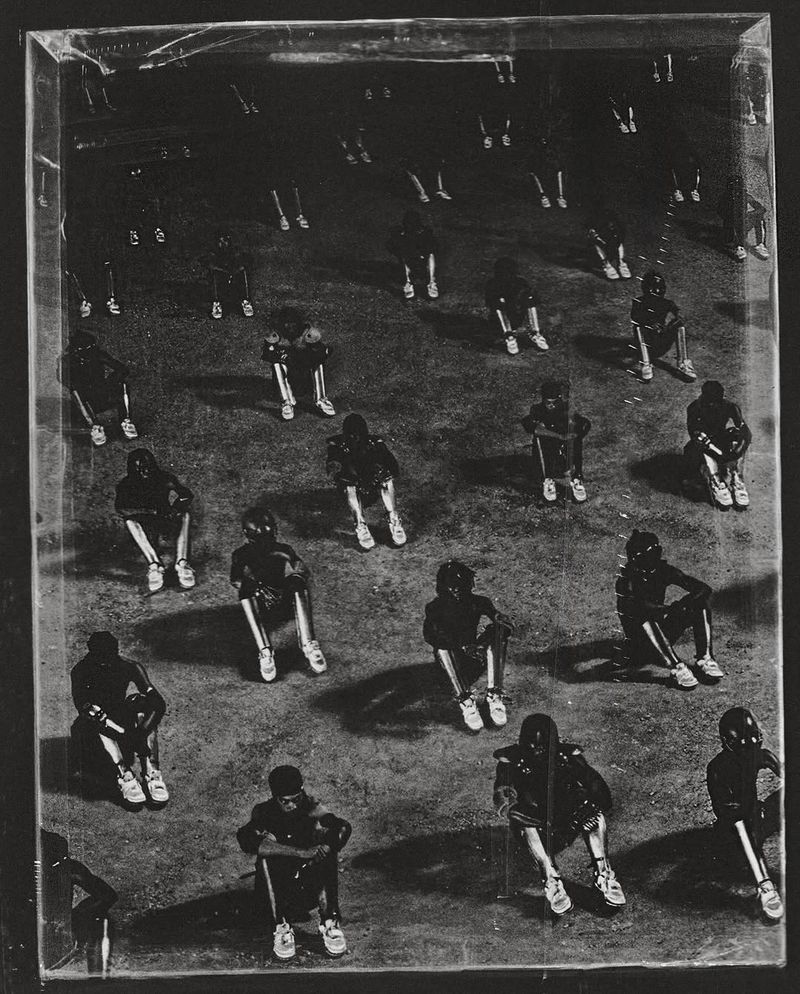 Monochromatic art piece featuring a large group of people in dark clothing sitting in rows, captured in a square composition.