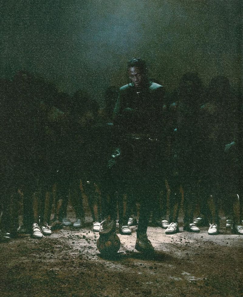 A dark image of an adult male football player stands with a ball with a team of shadowy figures behind him on a dark and unclear field.