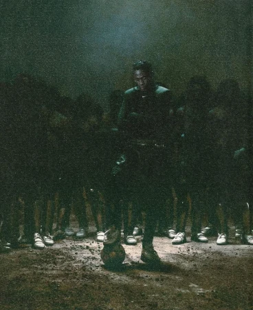 A dark image of an adult male football player stands with a ball with a team of shadowy figures behind him on a dark and unclear field.