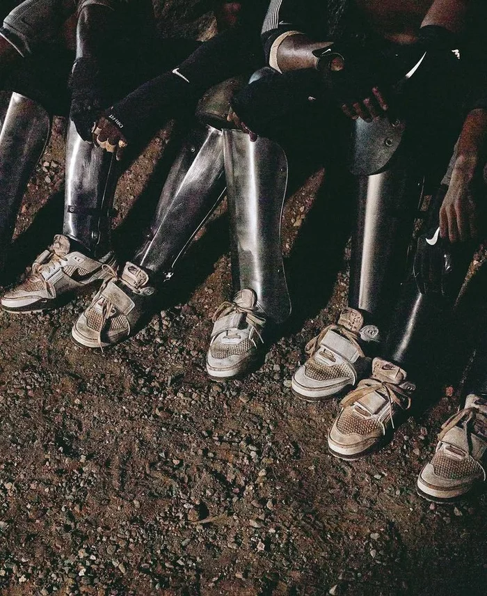 Close-up of several individuals wearing metallic shin guards and sneakers seated outdoors on rough dirt ground