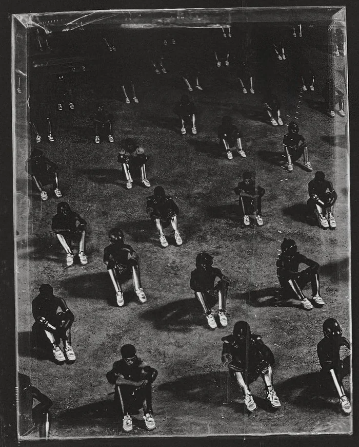 High-angle monochrome shot of multiple seated individuals arranged in a grid-like pattern on a textured ground.