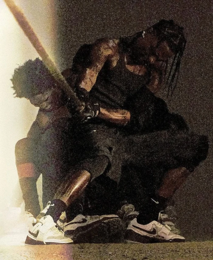 Travis Scott holding a bat in a grainy, low-lit portrait with a second figure crouching beside him
