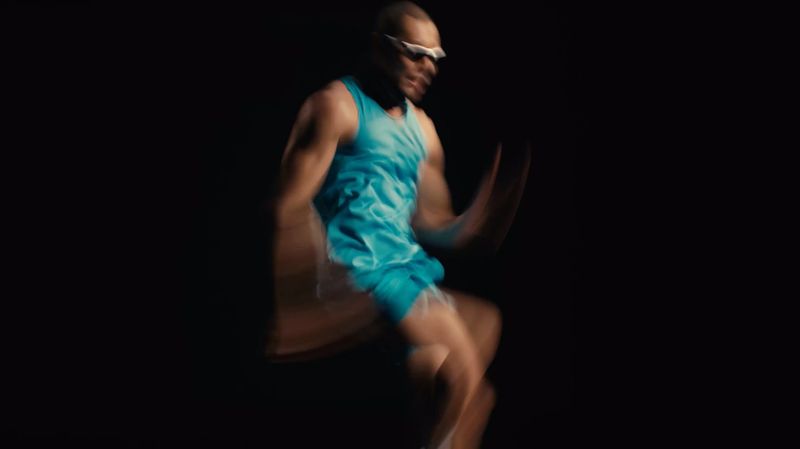 Man in blue athletic wear dynamically poses mid-motion against a stark black backdrop, embodying energy and fitness in a studio environment.
