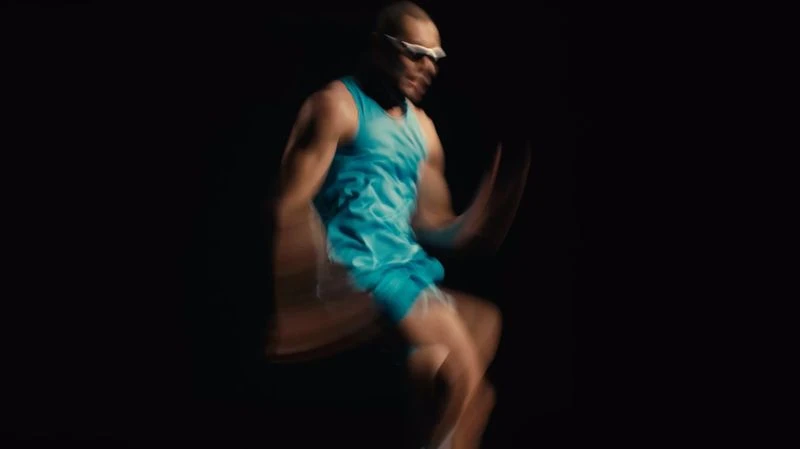 Man in blue athletic wear dynamically poses mid-motion against a stark black backdrop, embodying energy and fitness in a studio environment.
