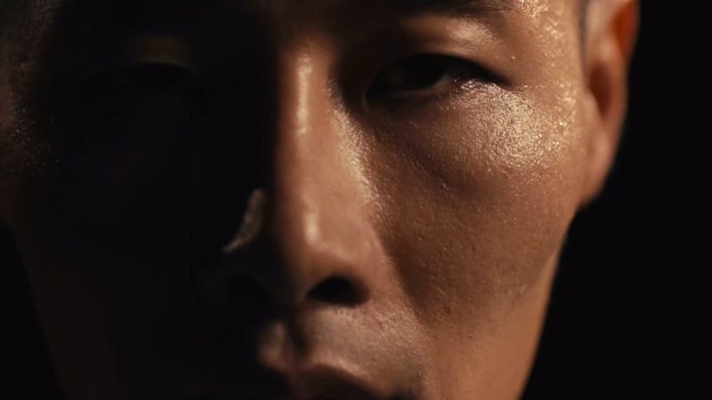 Close-up portrait of a sweaty adult male face in a dark studio, emphasizing skin texture, lighting, and the intense expression of the eyes.