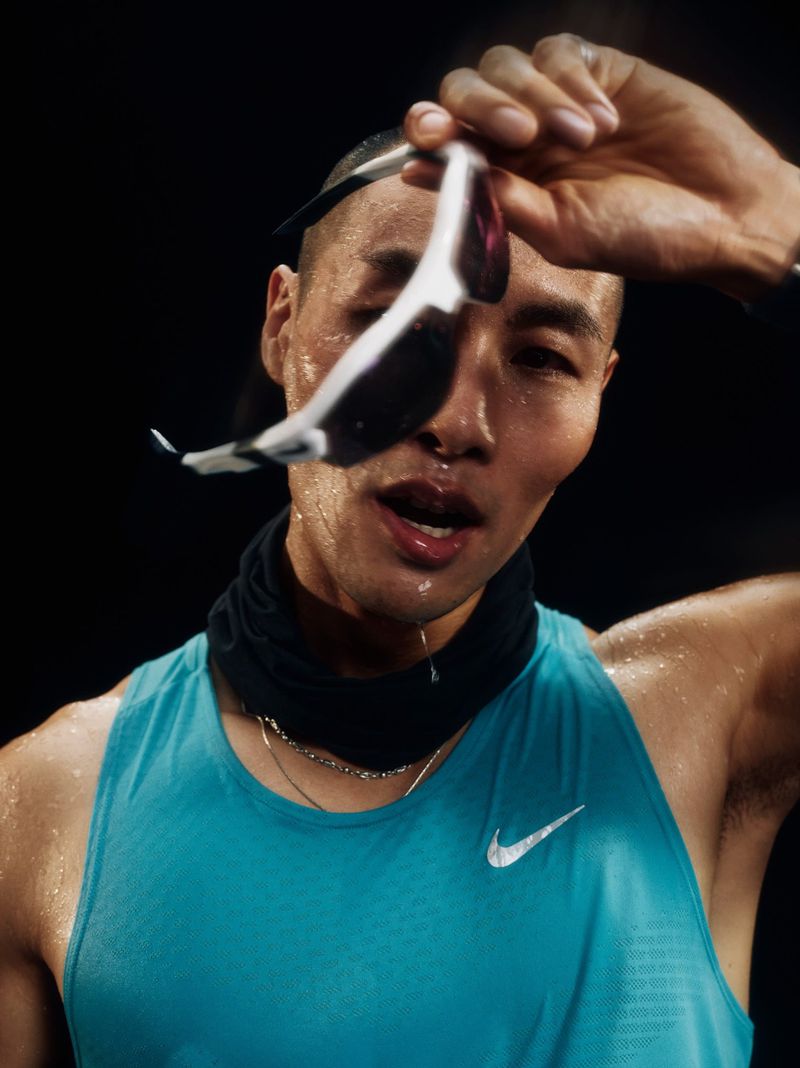 Close-up of a sweaty, athletic, Asian adult male wearing a turquoise tank top, black neck gaiter, holding sunglasses against a dark background.