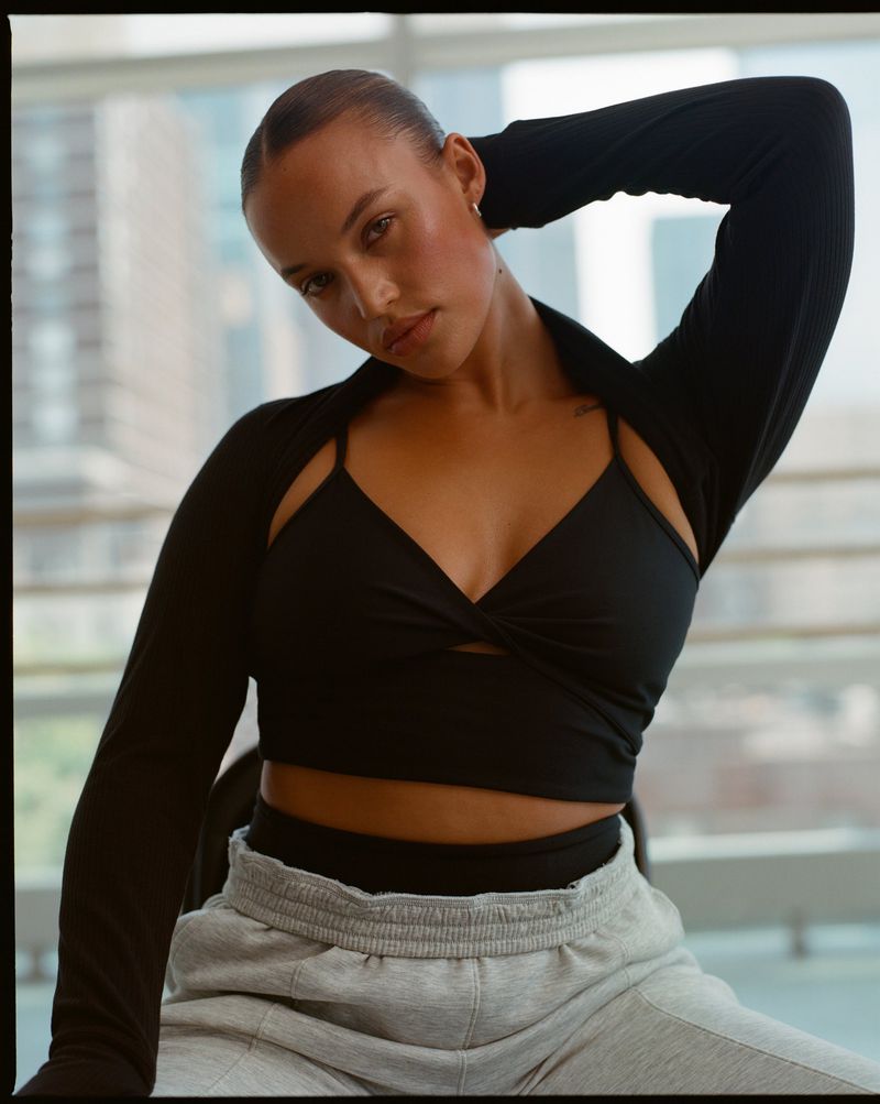 Young woman in athleisure wear poses in front of large window with cityscape background, wearing a black top and grey sweatpants.