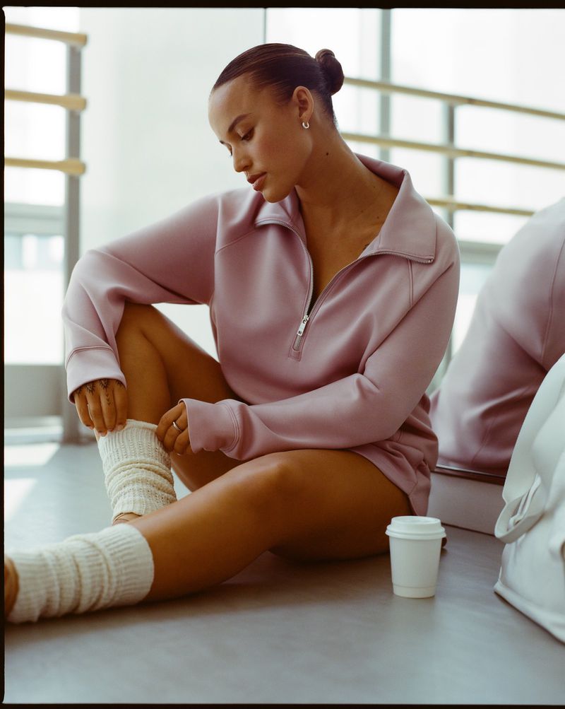 Young woman in athletic wear sits on floor wearing socks, pink quarter-zip, and hair in a bun inside a dance studio.