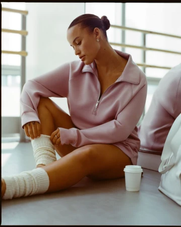 Young woman in athletic wear sits on floor wearing socks, pink quarter-zip, and hair in a bun inside a dance studio.