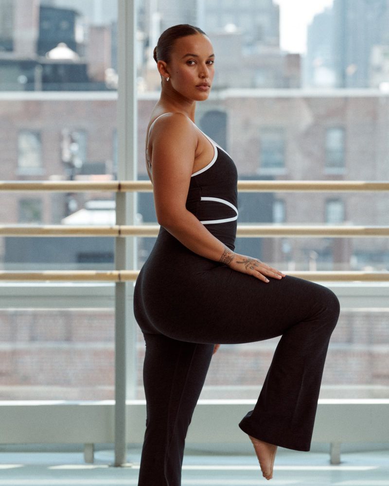 A woman in a black bodysuit with white trim poses in an indoor dance studio with a leg lifted against a barre, buildings visible outside.