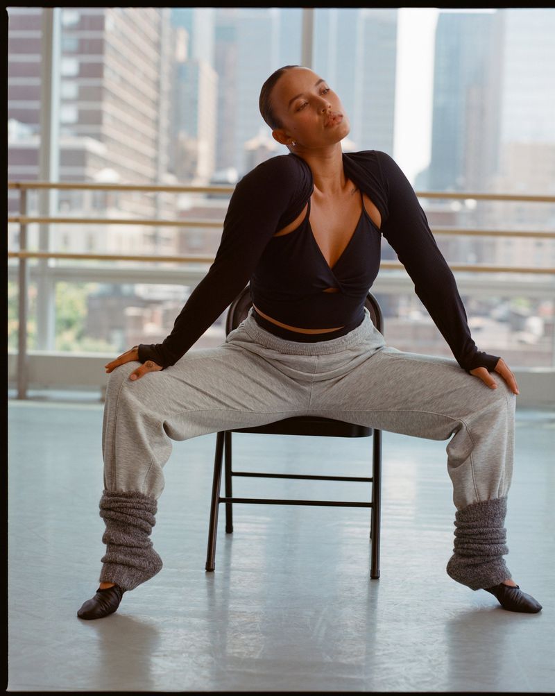 Young woman in ballet shoes and leg warmers sits on a chair in a dance studio with a cityscape view in the background.