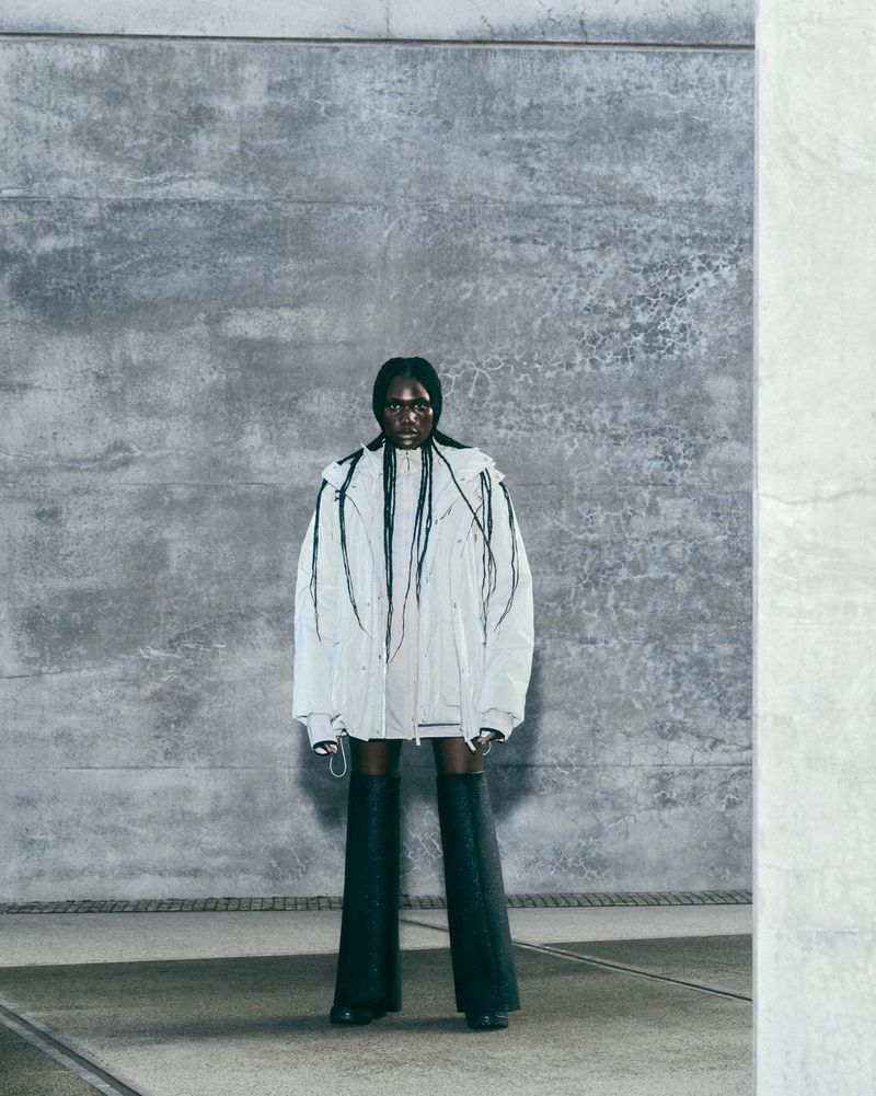 A full shot of an adult female with long braids, wearing a white jacket and thigh-high boots, stands against a concrete wall background.