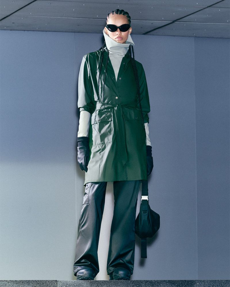Woman in green trench coat, black pants, and sunglasses posing on stairs in an urban setting. Fashion editorial photography.