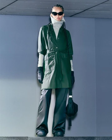 Woman in green trench coat, black pants, and sunglasses posing on stairs in an urban setting. Fashion editorial photography.