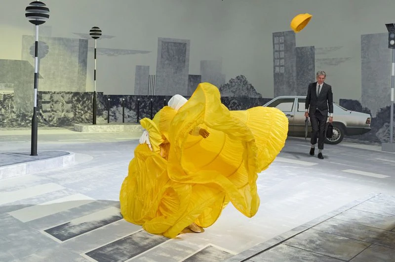 An abstract fashion shot in a studio featuring a person in a yellow costume on a grey crosswalk with a man in a suit walking in background.