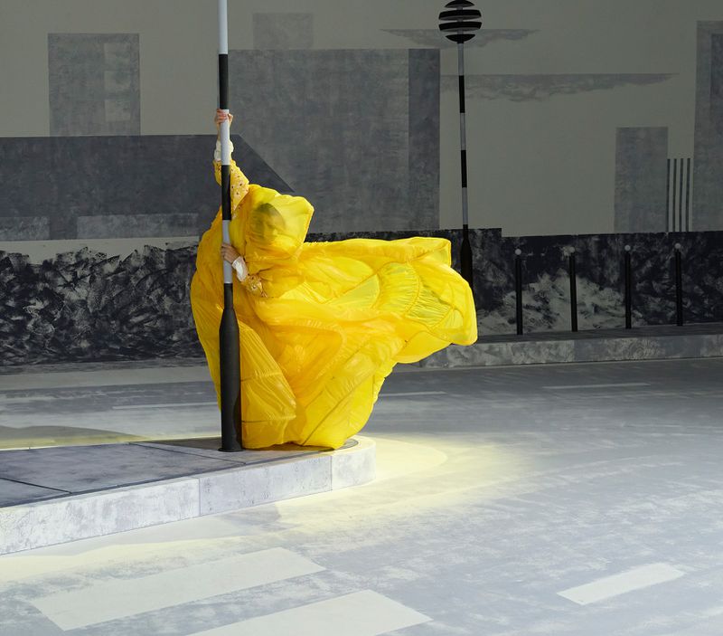 A person in a vibrant yellow costume clings to a lamp post on a city street with monochromatic buildings in the background.