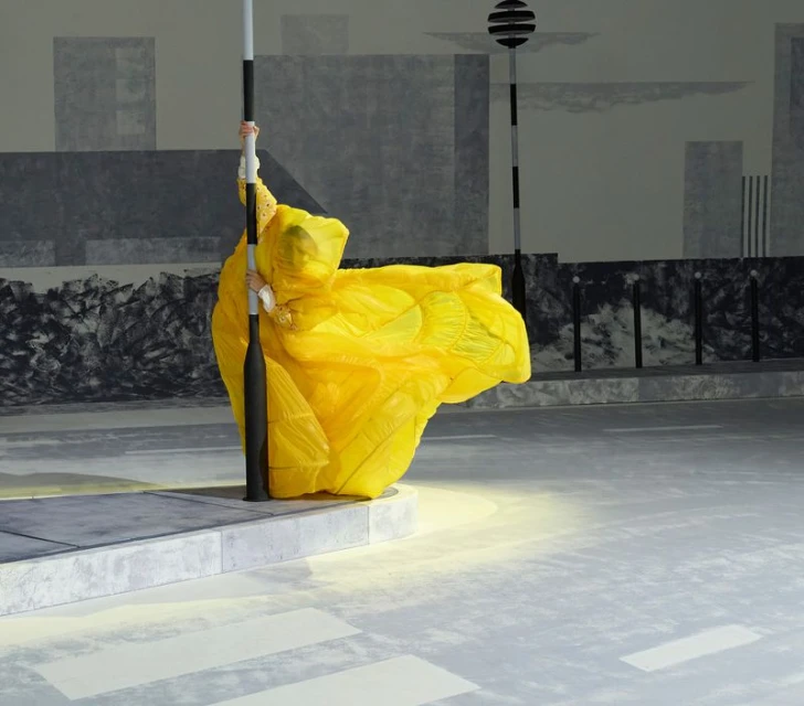 A person in a vibrant yellow costume clings to a lamp post on a city street with monochromatic buildings in the background.