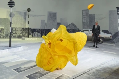 Figure in voluminous yellow ruffled dress in foreground with man walking in background on a surreal street set