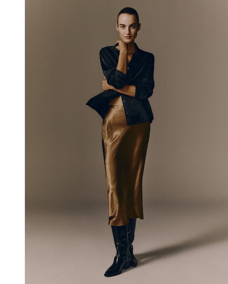 Fashion shot of a woman in a silky midi skirt, black jacket, and knee-high boots, posing in a studio with soft, neutral lighting.