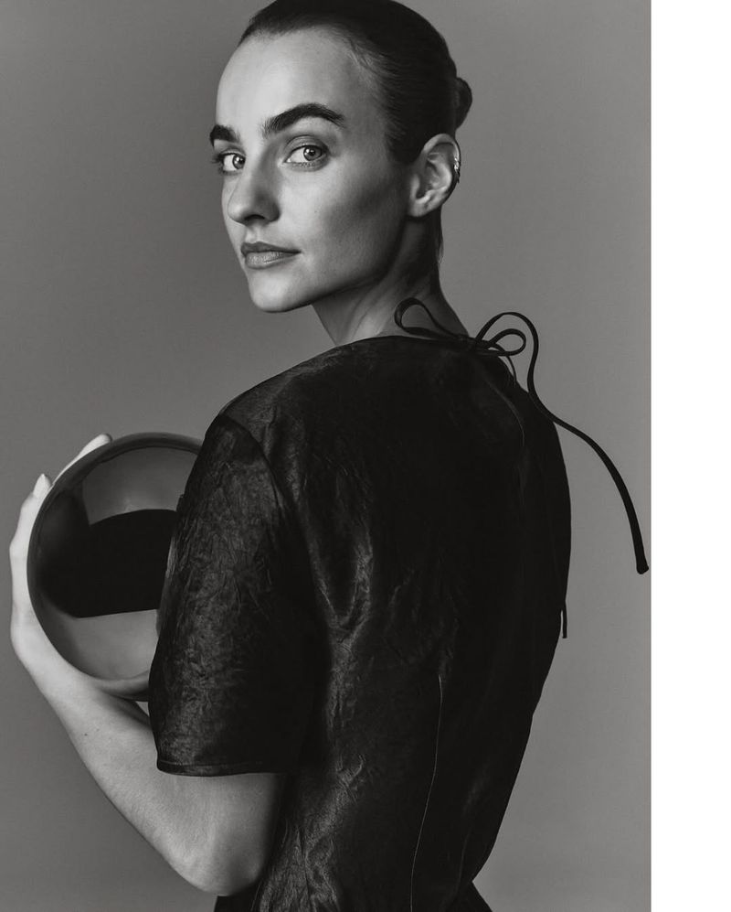Monochrome portrait of woman in dark top, gazing over shoulder while holding reflective ball. High-contrast, clean studio background.