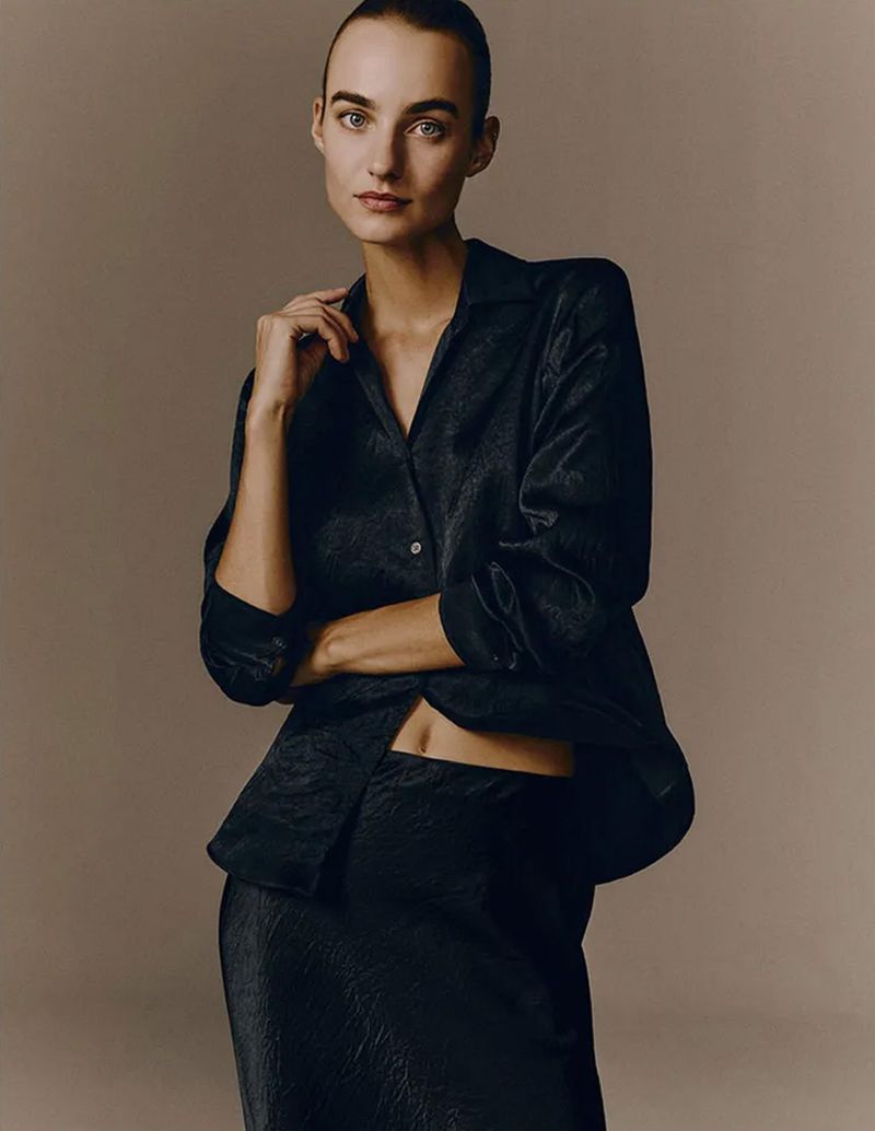 Woman in a dark, patterned silky shirt and skirt poses in a studio, exuding minimalist fashion and beauty against a neutral background.