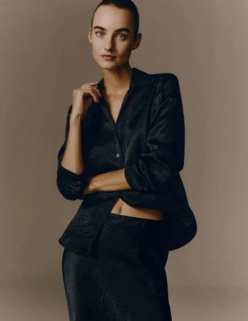 Woman in a dark, patterned silky shirt and skirt poses in a studio, exuding minimalist fashion and beauty against a neutral background.
