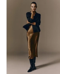 Model Maartje Verhoef wearing a dark silk shirt and golden midi skirt with tall black boots against a neutral grey studio background