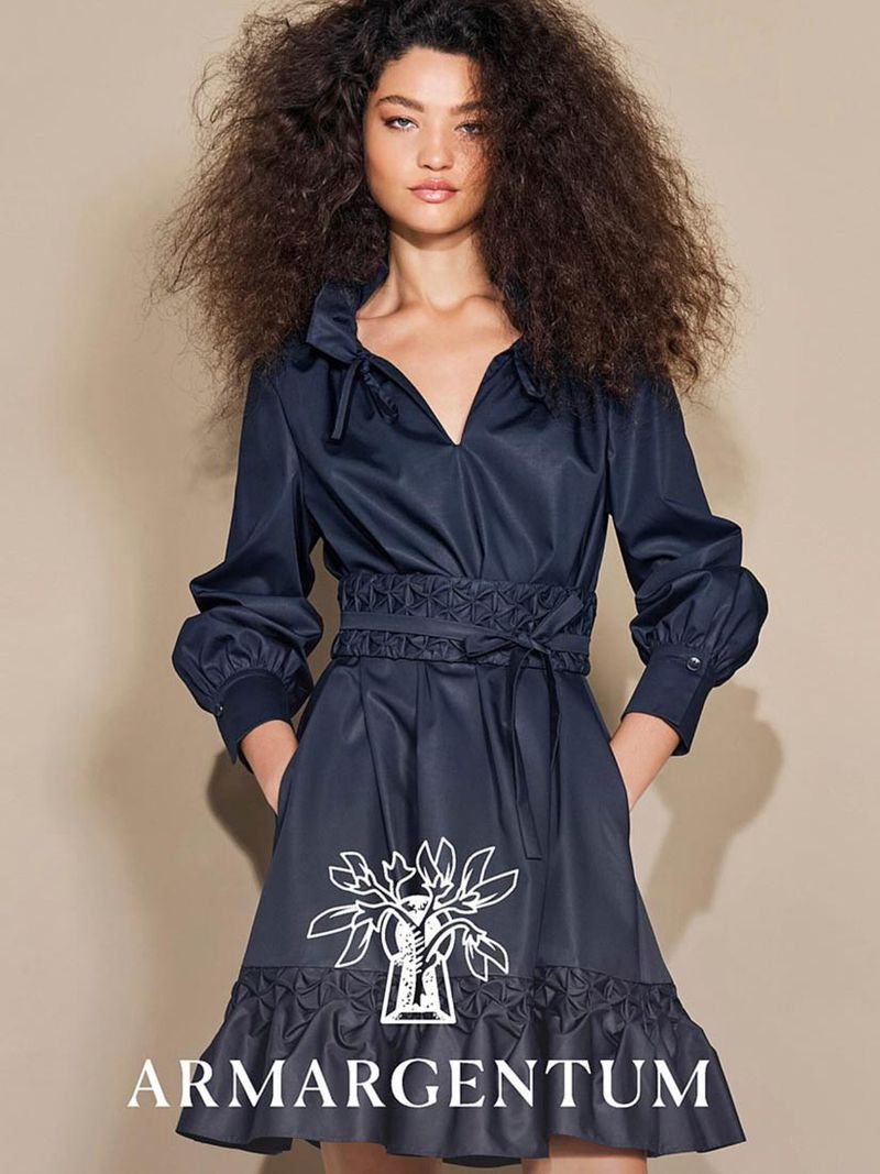 Fashion model wearing a dark blue collared dress with long sleeves, standing indoors against a neutral wall with soft lighting. 