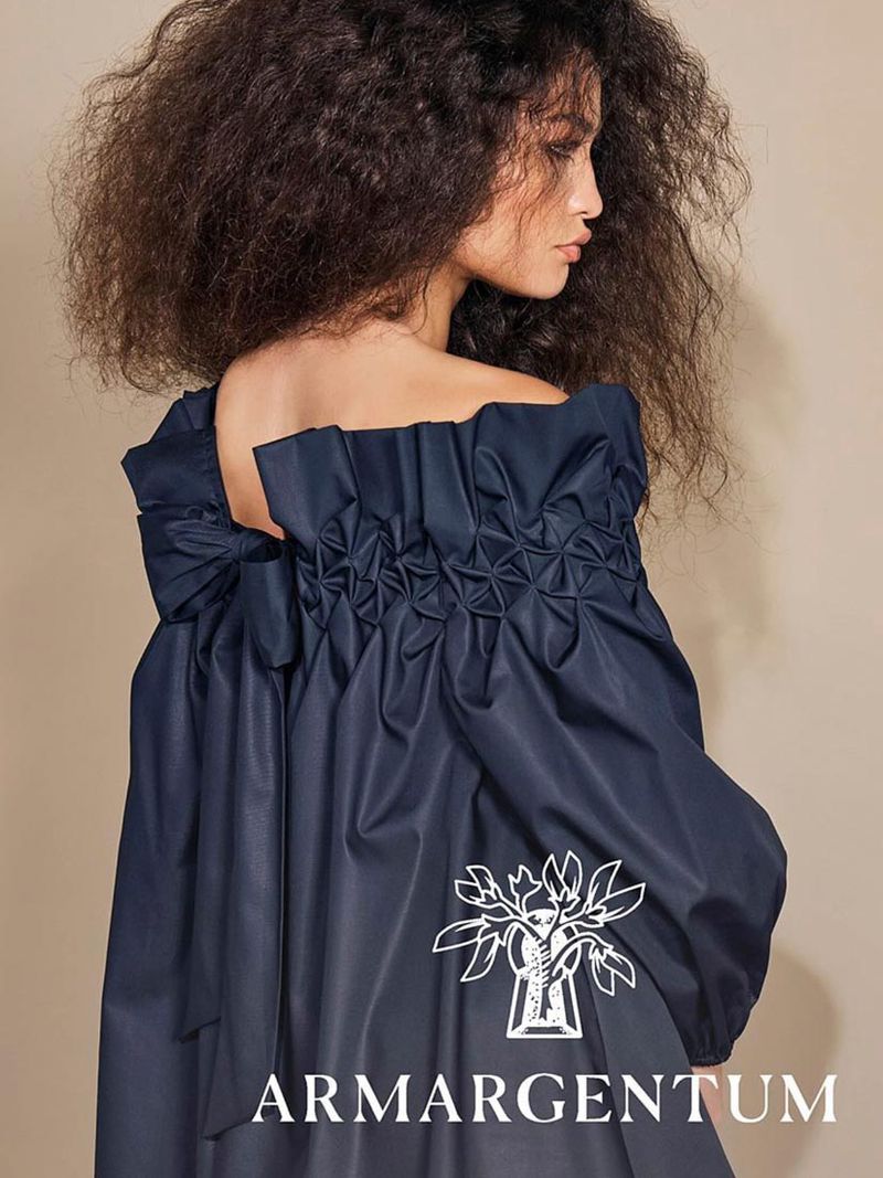 Brunette woman in off-shoulder dress with textured bodice and back bow. The dress is navy blue with a white logo on the skirt.