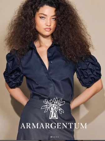 A stylish fashion portrait of a woman with curly dark hair, wearing a blue button-down shirt and dark skirt, showcasing a clothing brand.
