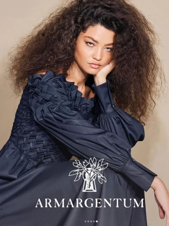Elegant woman with curly hair poses in a sophisticated navy dress against a tan backdrop, showcasing the brand Armargentum.
