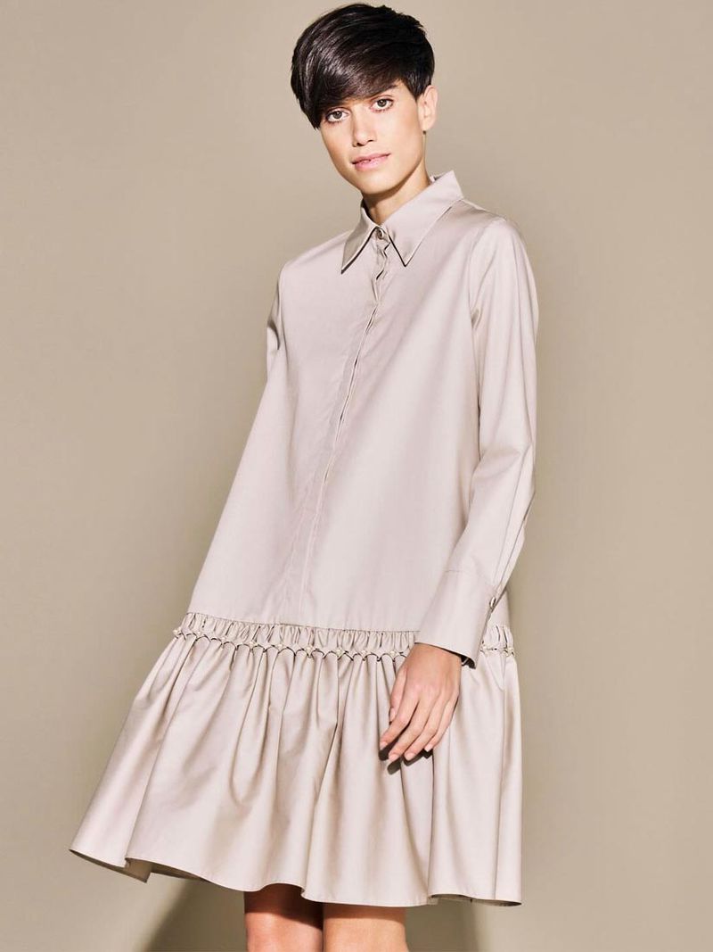 A woman models a neutral-toned, collared, long-sleeved mini dress with a ruffled hem against a matching solid background in a fashion studio shot.