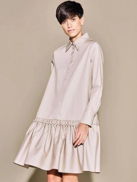 A woman models a neutral-toned, collared, long-sleeved mini dress with a ruffled hem against a matching solid background in a fashion studio shot.