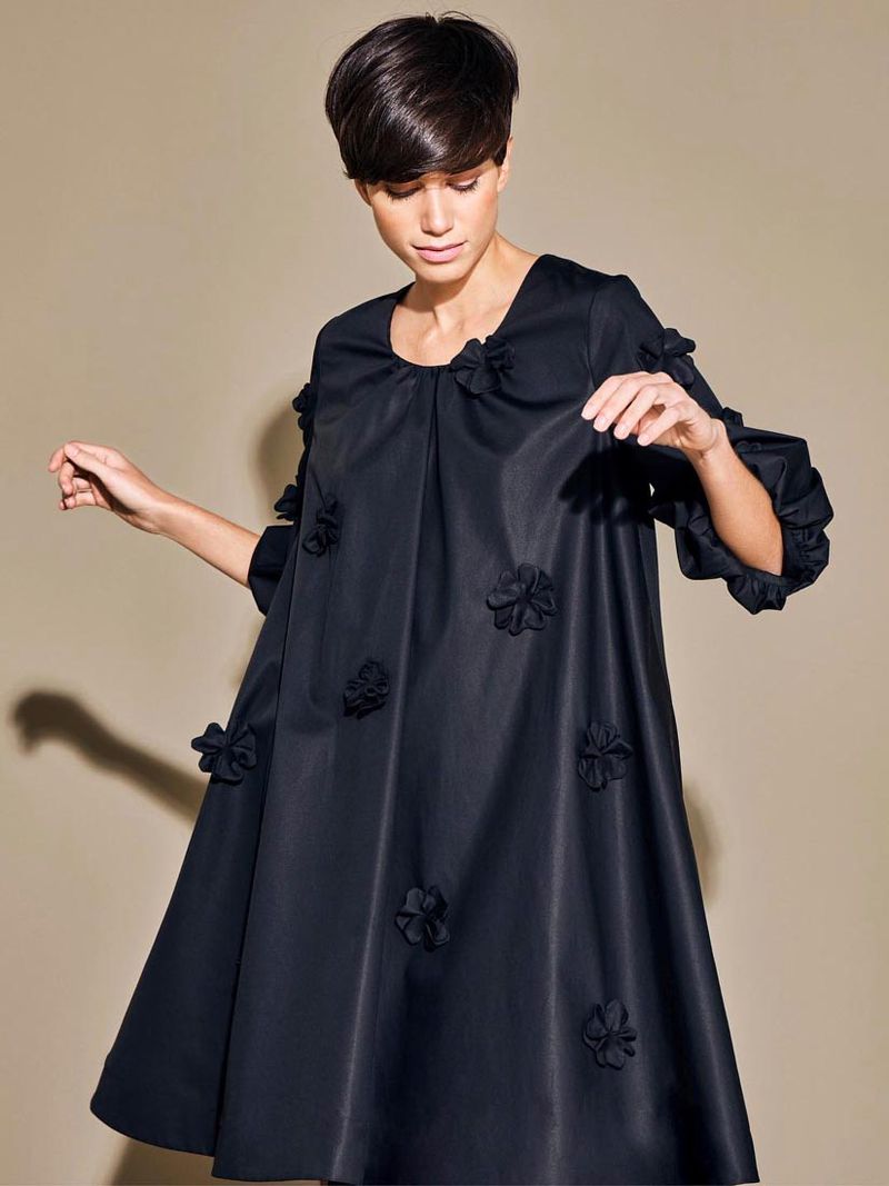 Fashionable woman in a short dark dress adorned with floral appliques posing against a neutral backdrop in a studio setting.